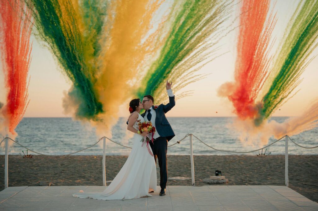 Braut und Bräutigam küssen sich am Strand vor dem Hintergrund eines farbenprächtigen Feuerwerks bei Sonnenuntergang.