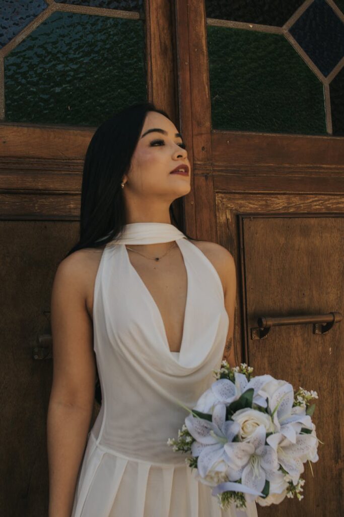 Bride in white dress holding flower bouquet against wooden backdrop.