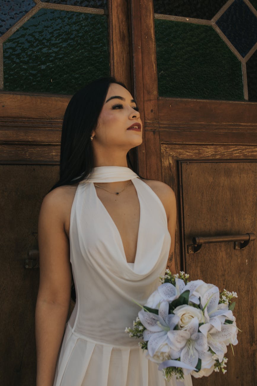 Bride in white dress holding flower bouquet against wooden backdrop.