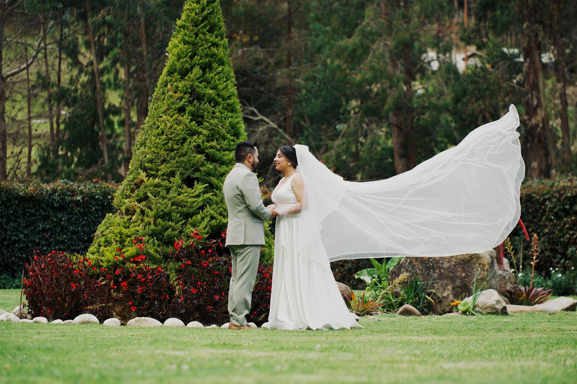 Braut und Bräutigam stehen mit wehendem Schleier im Garten. Grün und Blumen umgeben sie.